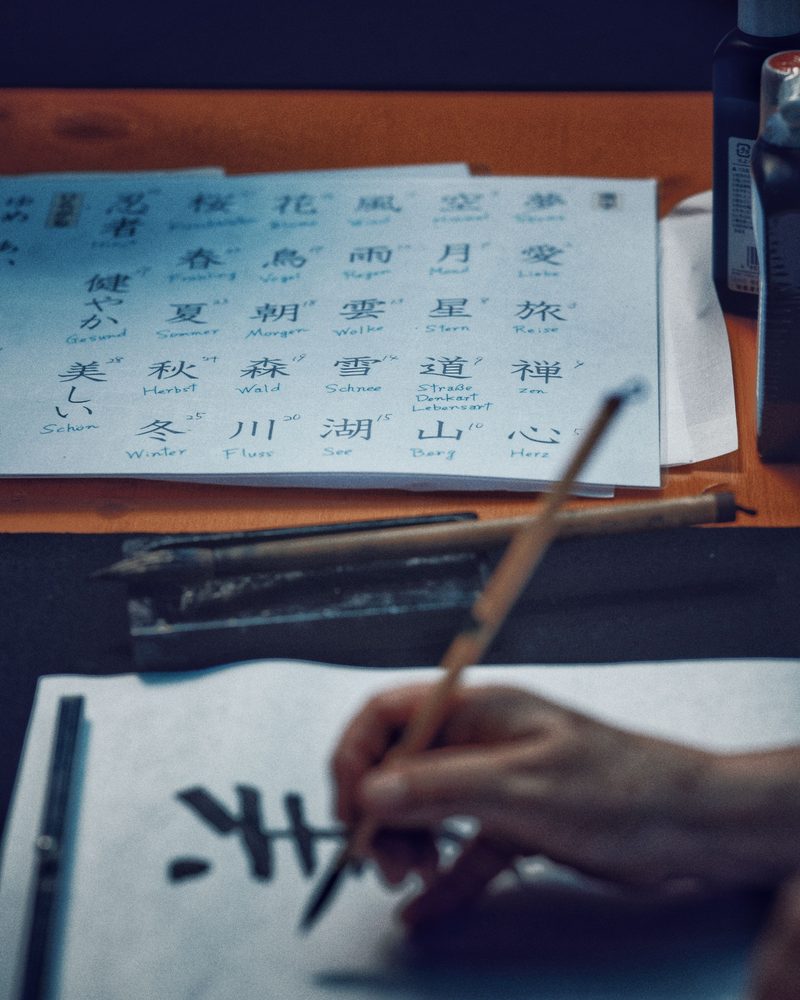 Atelier de calligraphie : une main trace un caractère au pinceau, à côté d’une fiche de kanji annotée et de flacons d’encre.