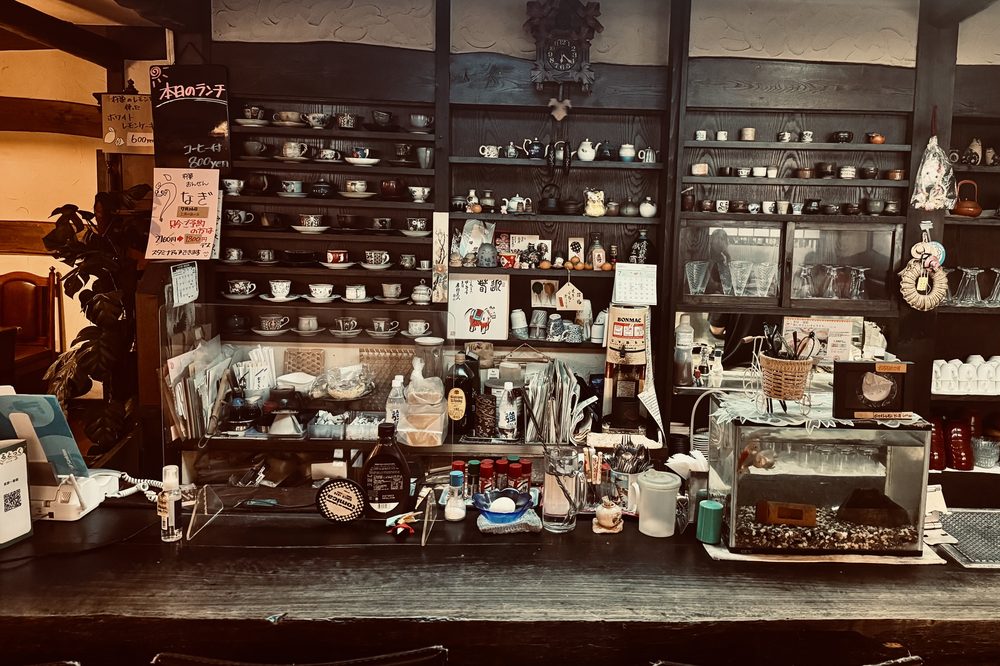 Comptoir d’un vieux café japonais : tasses alignées sur des étagères de bois sombre, menus écrits à la main, horloge à coucou.